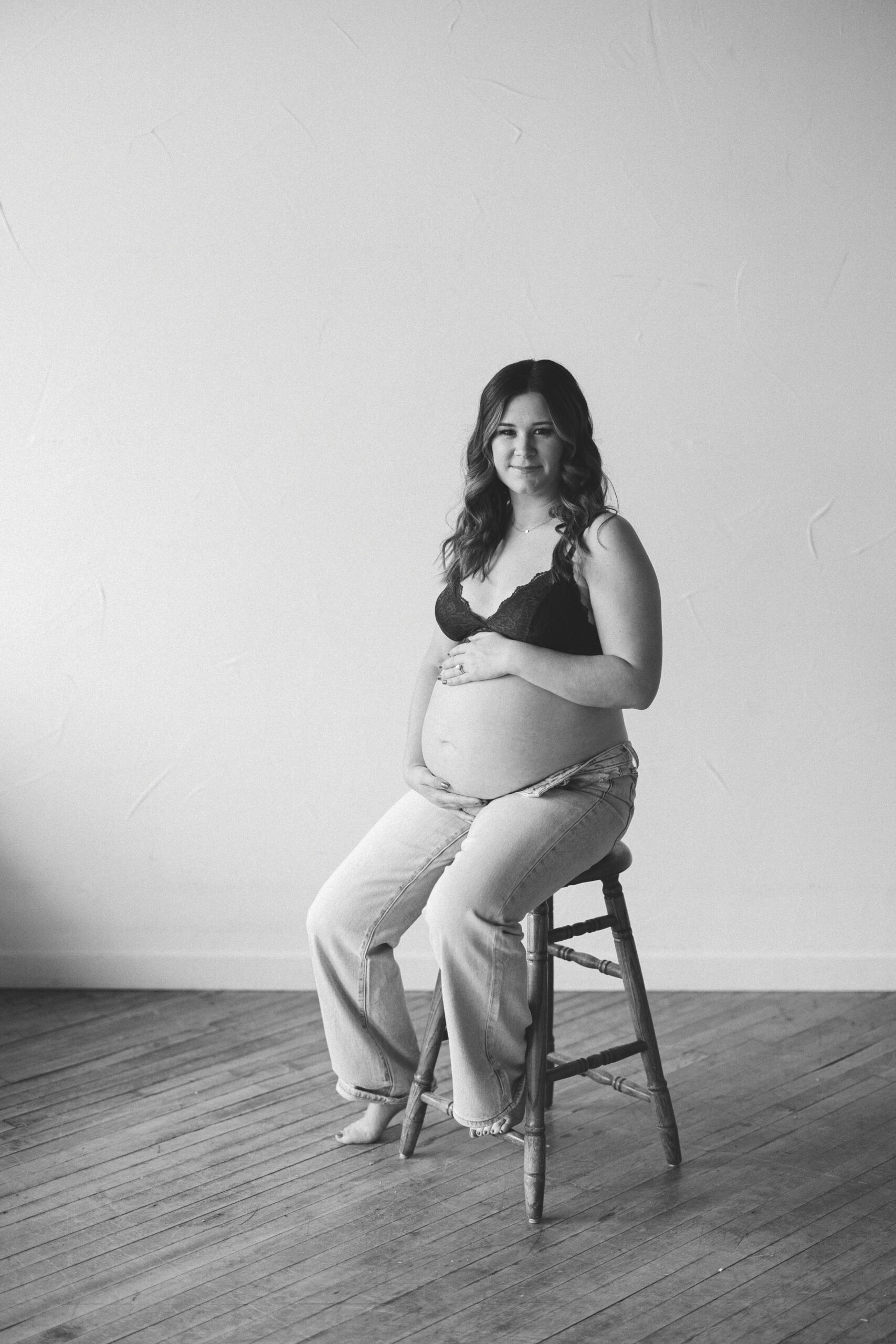 Pregnant mother sitting on a stool, looking effortlessly beautiful in jeans and a bralette during a Dallas Maternity session.